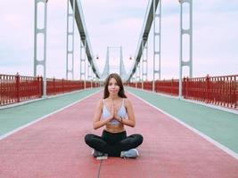 beautiful, sporty, fit, young caucasian woman in sportswear and sneakers sitting in lotus pose on the bridge. Sport, activity, yoga, urban lifestyle concept photo