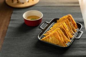 Easy recipe, snacks, minced pork meat filled fried wontons sort in black dish on grey placemat, brown wood table served with sweet plum sauce in red bowl photo