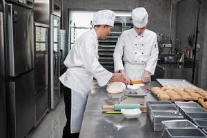 dos chefs asiáticos profesionales con uniformes y delantales de cocinero blanco están amasando masa de pastelería y huevos, preparando pan y comida fresca de panadería, horneando en el horno en la cocina de acero inoxidable del restaurante. foto