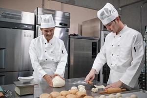 Two professional Asian male chefs in white cook uniforms and aprons are kneading pastry dough and eggs, preparing bread and fresh bakery food, baking in oven at stainless steel kitchen of restaurant. photo