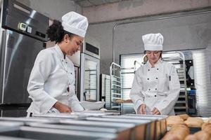 Two professional female chefs in white cook uniforms and aprons knead pastry dough and eggs, prepare bread, cookies, and fresh bakery food, baking in oven at a stainless steel kitchen of a restaurant. photo