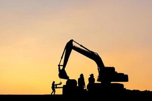 Silhouette of Excavator and worker team at construction site with blurred sunset background