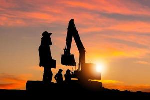 silueta del capataz y el equipo de trabajadores en el sitio de construcción, trabajador de la construcción de carreteras y excavadora con fondo borroso de puesta de sol