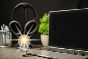 A bright light bulb and laptop are black blank screen on work table. Concept of a startup that drives the economy, finance, banking. Wireless headphones in the working room. Blurred, black background