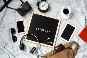 Saturday flat lay concept with multimedia entertainment accessories, sunglasses, alarm clock, gadget, camera, headphone and coffee for refreshment on white background