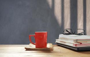 taza de café roja y anteojos en libros apilados, laptop negra en una mesa de madera frente a una pared de cemento en el salón