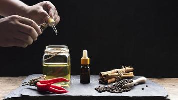 Man's hand holding pipette, dropper bottle with various herbs and herbal essential oil in glass jar on black stone plate in vintage dark tone style, selective focus photo