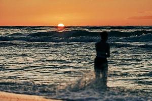 Female silhouette in blue sea waves at summer sunset, half sun below horizon, beachfront holiday
