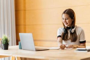 Happy asian businesswoman wearing headset make notes talk by conference call doing video chat on laptop in office, smiling chinese support service agent write information consult customer photo