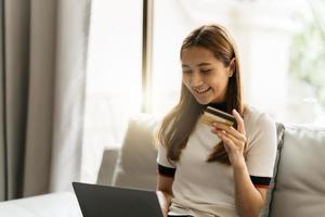 Online shopping concept. Dreamy asian woman with computer laptop and credit card sitting on couch, thinking about internet purchases or food delivery, free space. photo