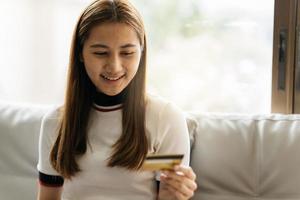 Online shopping concept. Dreamy asian woman with computer laptop and credit card sitting on couch, thinking about internet purchases or food delivery, free space. photo