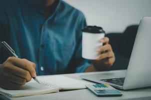 Male hand holding pen and writing at notebook and coffee cup on desk with notebook computer and calculator. photo