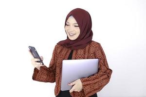 Young Asian Islam woman is standing and smiling face when looking on the phone with holding laptop photo