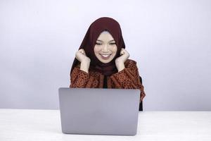 Young Asian Islam woman wearing headscarf is shocked and excited with what she see on laptop on the table. photo