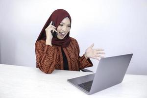Young Asian Islam woman wearing headscarf is smiling and excited with when she call on the phone and front on the laptop on the table. photo