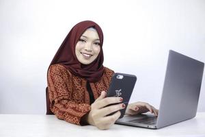 una joven musulmana asiática con pañuelo en la cabeza sonríe en un teléfono móvil con una laptop en la mesa. foto