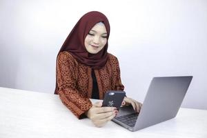 Young Asian Islam woman wearing headscarf is smiling on a mobile phone with laptop on the table. photo