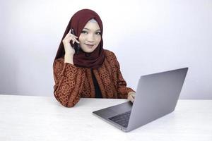 Young Asian Islam woman wearing headscarf is smiling and excited with when she call on the phone and front on the laptop on the table. photo