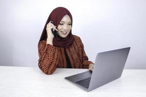 Young Asian Islam woman wearing headscarf is smiling and excited with when she call on the phone and front on the laptop on the table. photo