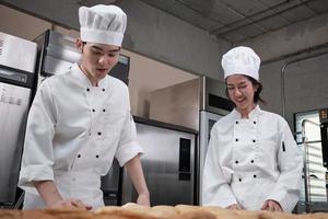 Two professional Asian chefs in white cook uniforms and aprons knead pastry dough and eggs, prepare bread, cookies, and fresh bakery food, baking in an oven in a restaurant stainless steel kitchen. photo