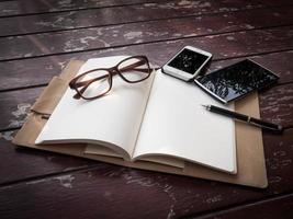 espacio de trabajo con gafas, bolígrafo y teléfono inteligente, papel de notas y cuaderno en una mesa de madera antigua foto