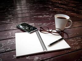 espacio de trabajo con gafas, bolígrafo, teléfono inteligente y taza de café, papel de notas y cuaderno en una mesa de madera antigua foto