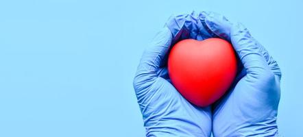 Both hands were wearing gloves and holding a red heart. Concept world heart foundation organizes world heart day, copy space on left for design or text, close-up, blurred blue background photo