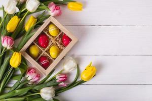 Top view of colorful easter eggs on a bed of straw in a long wooden box on a white wooden table and tulips, place for text photo