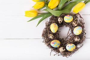 Beautiful yellow tulips with colorful quail and chicken eggs in wreath on white wooden background. Spring and Easter holiday concept. photo