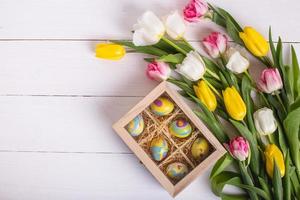 Top view of colorful easter eggs on a bed of straw in a long wooden box on a white wooden table and tulips, place for text photo