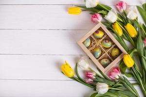 Top view of colorful easter eggs on a bed of straw in a long wooden box on a white wooden table and tulips, place for text photo