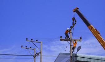 Two electricians with crane truck are installing electrical equipment on power pole against blue sky background