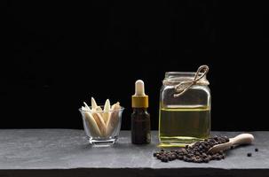 frasco cuentagotas con aceite esencial de hierbas en frasco de vidrio y rodajas de hierba de limón en un vaso pequeño con granos de pimienta negra en cuchara de madera sobre placa de pizarra negra en fondo oscuro foto