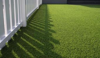 Sunlight and shadow of white wooden fence on green artificial turf surface in front yard of home, selective focus with copy space photo