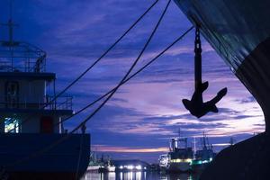 Cuerda de amarre de silueta con ancla de buques de carga atracados en el puerto a orillas del río contra el fondo del cielo crepuscular