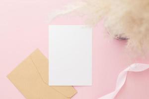 maqueta de invitación de tarjeta de felicitación en blanco 5x7 en sobre con flores de gypsophila y cinta sobre fondo de papel rosa pastel, capa plana, maqueta