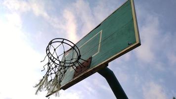 Vista frontal en ángulo bajo del viejo aro de baloncesto verde tenue y red rota con un fondo azul del cielo matutino en el campo deportivo público. foto