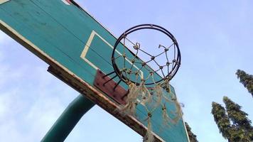 Vista frontal en ángulo bajo del antiguo aro de baloncesto verde tenue y red rota con un fondo azul del cielo matutino en el campo deportivo público. foto