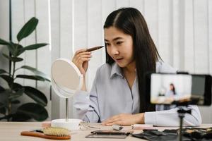 una joven asiática que hace vlogs sobre productos cosméticos para el cuidado de la piel en la mesa con su cámara de video y demuestra el uso de productos y reseñas para su canal de blog en línea. foto