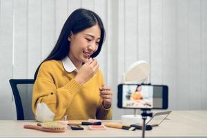 Young asian woman vlogging about cosmetics skin care items products on table with her video camera and demonstrates product use and reviews for her online blog channel. photo