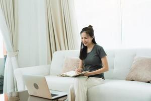 Asian woman sits on the sofa while taking notes from her computer screen while working or studying online at home. photo