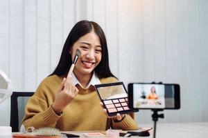 Beautiful asian woman blogger using camera phone recording vlog video live streaming and showing how to make up cosmetics at home. photo