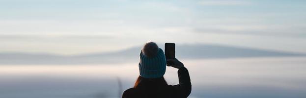 Adult woman use smartphone take a photo. Misty sunrise mountain banner background. photo