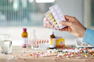 Hand holding medicines pill pack with colorful drugs spread on wooden table in the room background photo