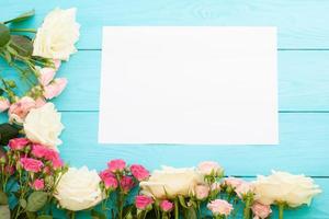 Frame of colorful roses on blue wooden background with paper card and copy space. Top view and selective focus. Mock up photo