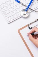 Female doctor writing a medical record case over clipboard on white working table with stethoscope, computer keyboard. Top view, flat lay, copy space photo