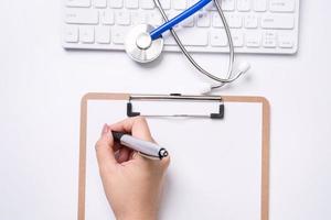 Female doctor writing a medical record case over clipboard on white working table with stethoscope, computer keyboard. Top view, flat lay, copy space photo