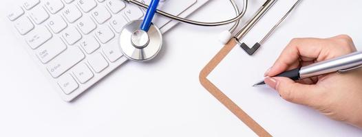 Female doctor writing a medical record case over clipboard on white working table with stethoscope, computer keyboard. Top view, flat lay, copy space photo