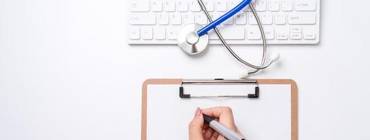 Female doctor writing a medical record case over clipboard on white working table with stethoscope, computer keyboard. Top view, flat lay, copy space photo