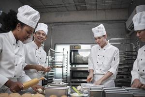 equipo gourmet profesional multirracial, cuatro chefs con uniformes de cocina blancos y delantales amasan masa de pastelería y harina, preparan pan y comida de panadería, hornean en el horno en la cocina del restaurante de acero inoxidable. foto